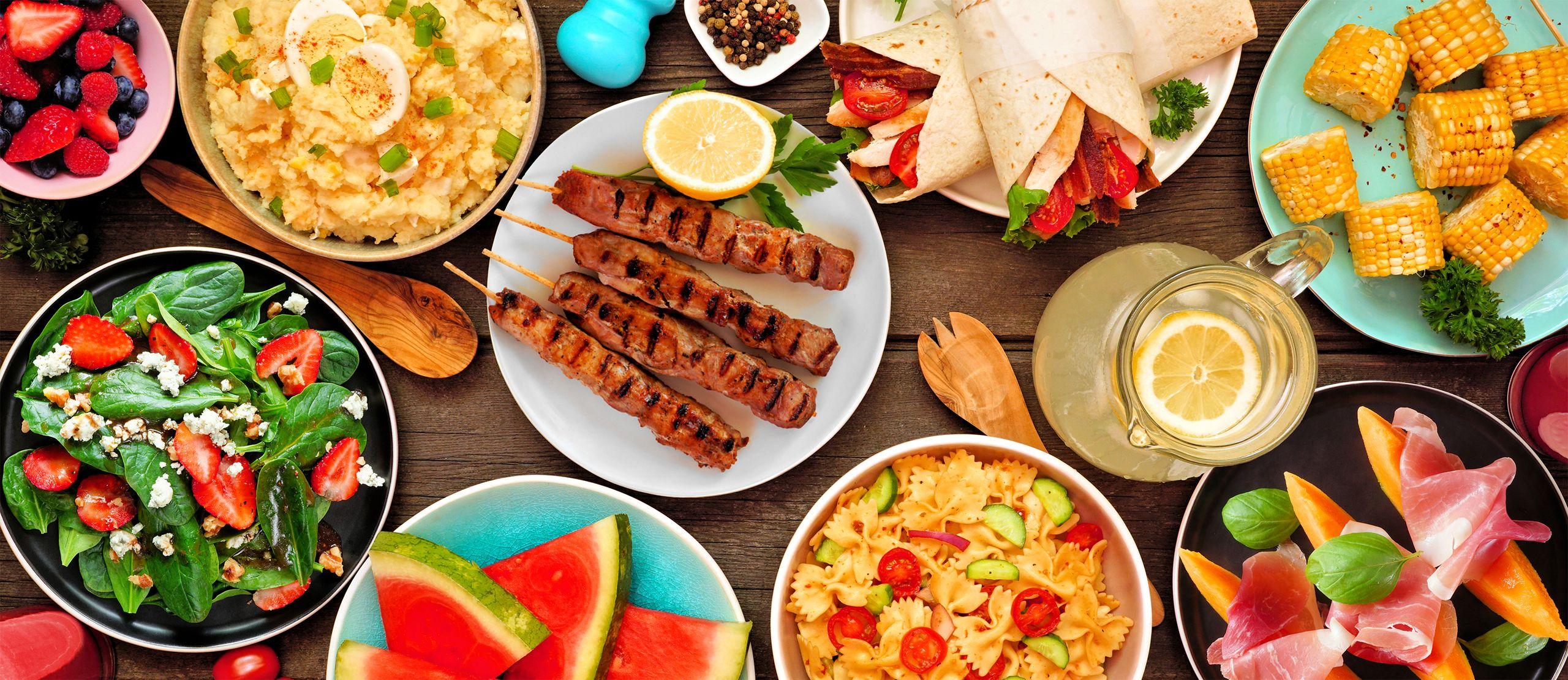 Overhead picnic spread: grilled skewers, spinach-strawberry salad, pasta salad, corn, watermelon, wraps, prosciutto-wrapped melon and lemonade.
