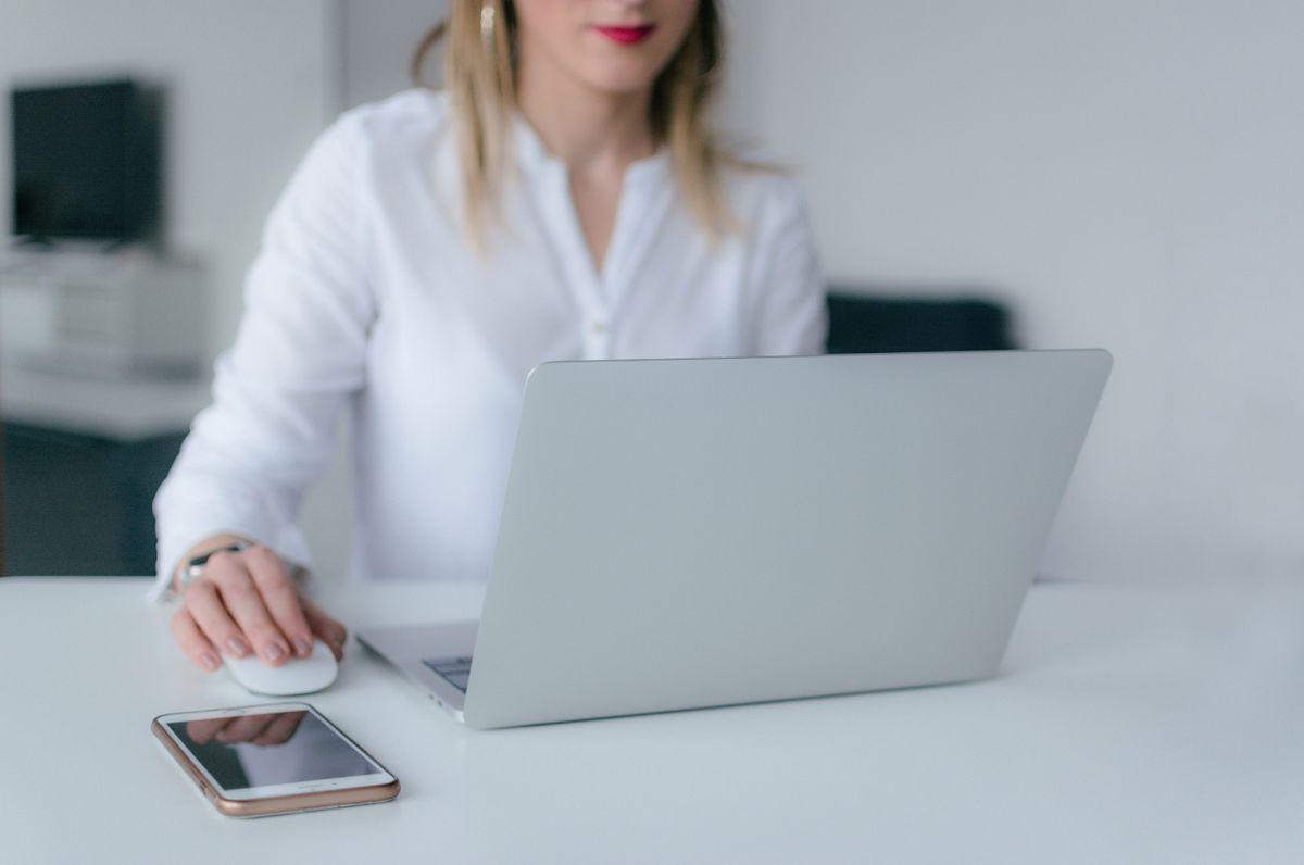 Woman in a white blouse using a laptop and mouse at a clean white desk with a smartphone beside her.